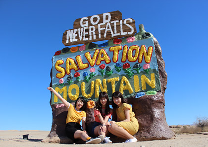 カリフォルニアの人気スポット、サルベーションマウンテンとデザートヒルズアウトレットを巡るツアーより。“GOD NEVER FAILS”の文字が印象的な入り口サインの前で笑顔で座る女性3人のツアー参加者